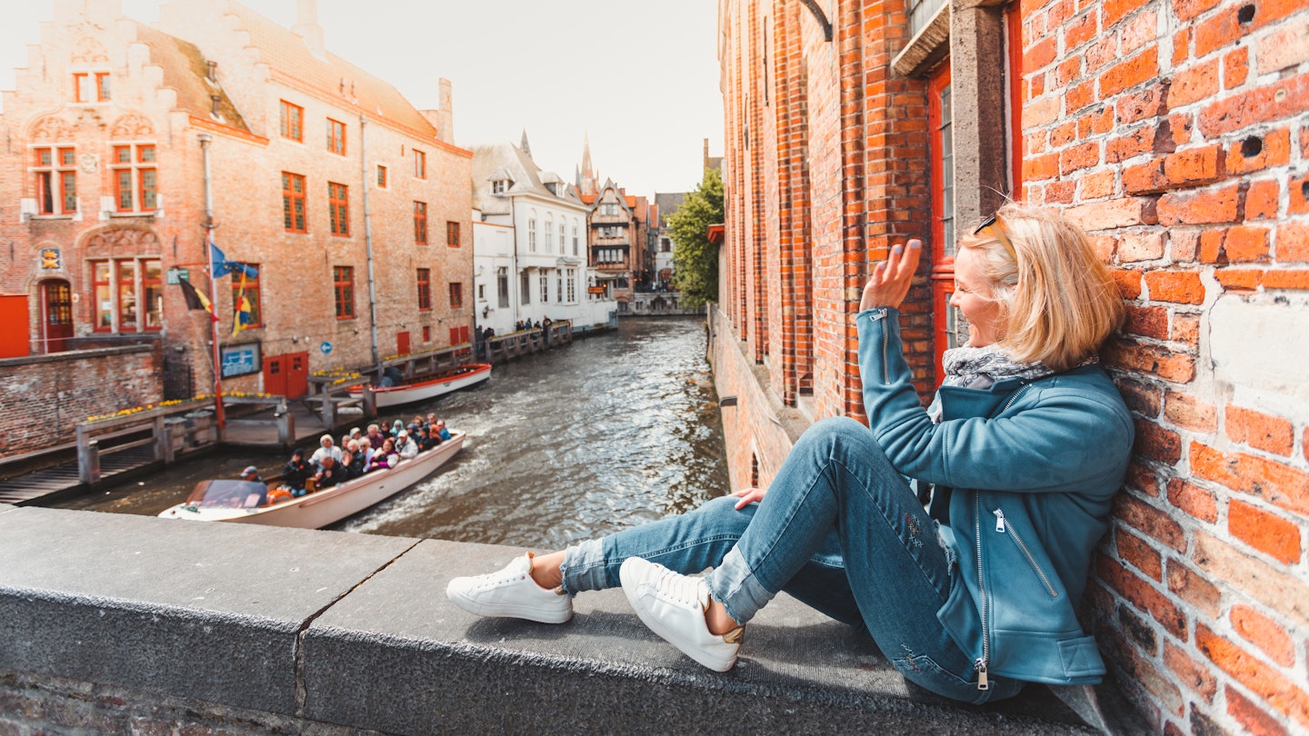 Young woman tourist sits on a bridge in the old city in Bruges in the morning in Belgium and waves a hand passing by a tourist boat with tourists.; Shutterstock ID 1400041118; your: Claire Naylor; gl: 65050; netsuite: Online editorial; full: Bruges things to do