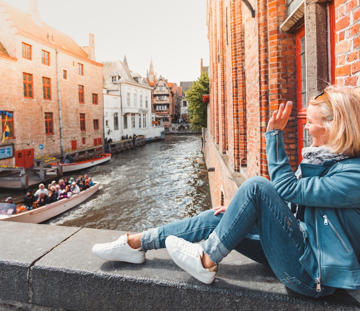 Young woman tourist sits on a bridge in the old city in Bruges in the morning in Belgium and waves a hand passing by a tourist boat with tourists.; Shutterstock ID 1400041118; your: Claire Naylor; gl: 65050; netsuite: Online editorial; full: Bruges things to do