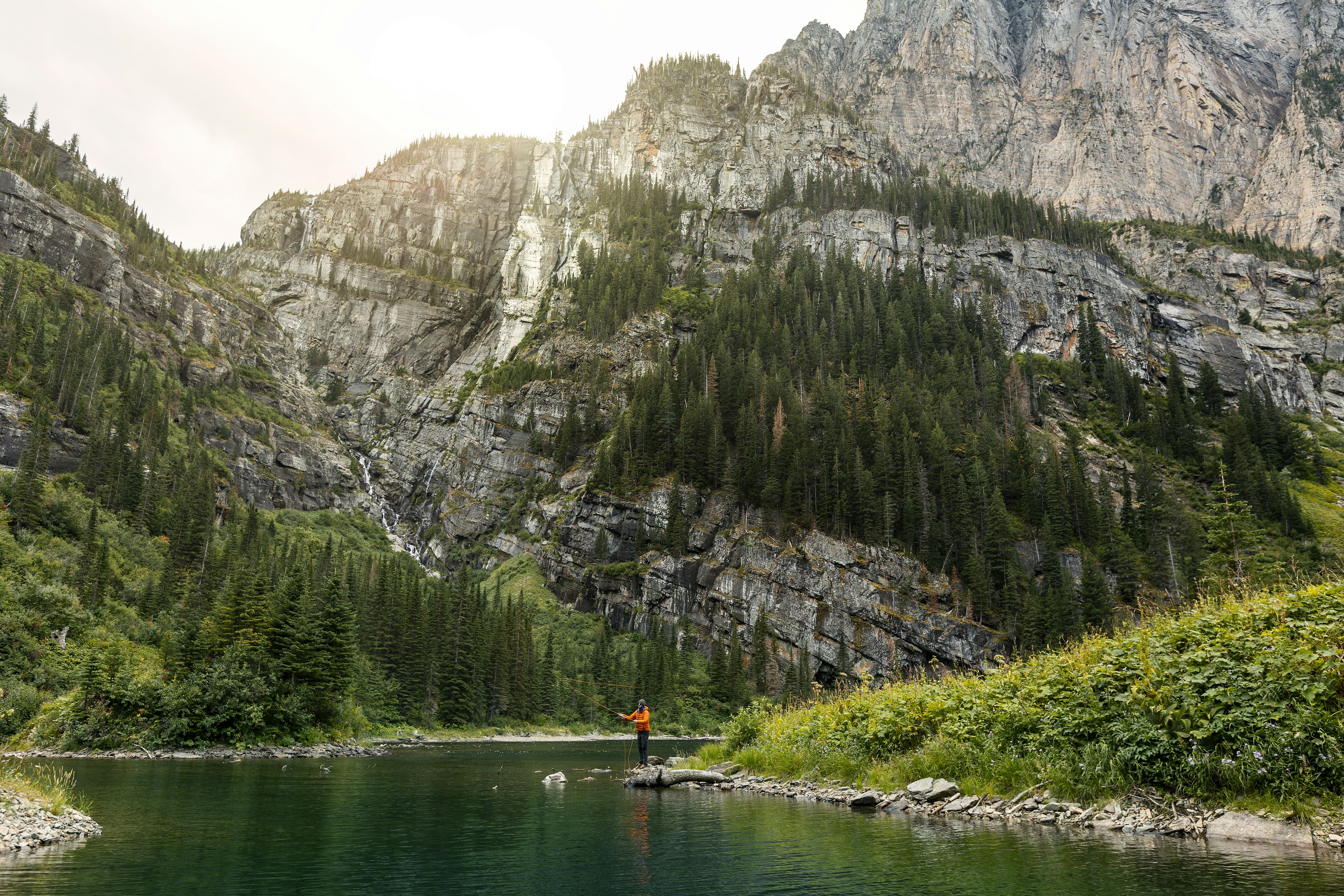 A fisher flings out a line in a river in the shadow of a vast granite cliff