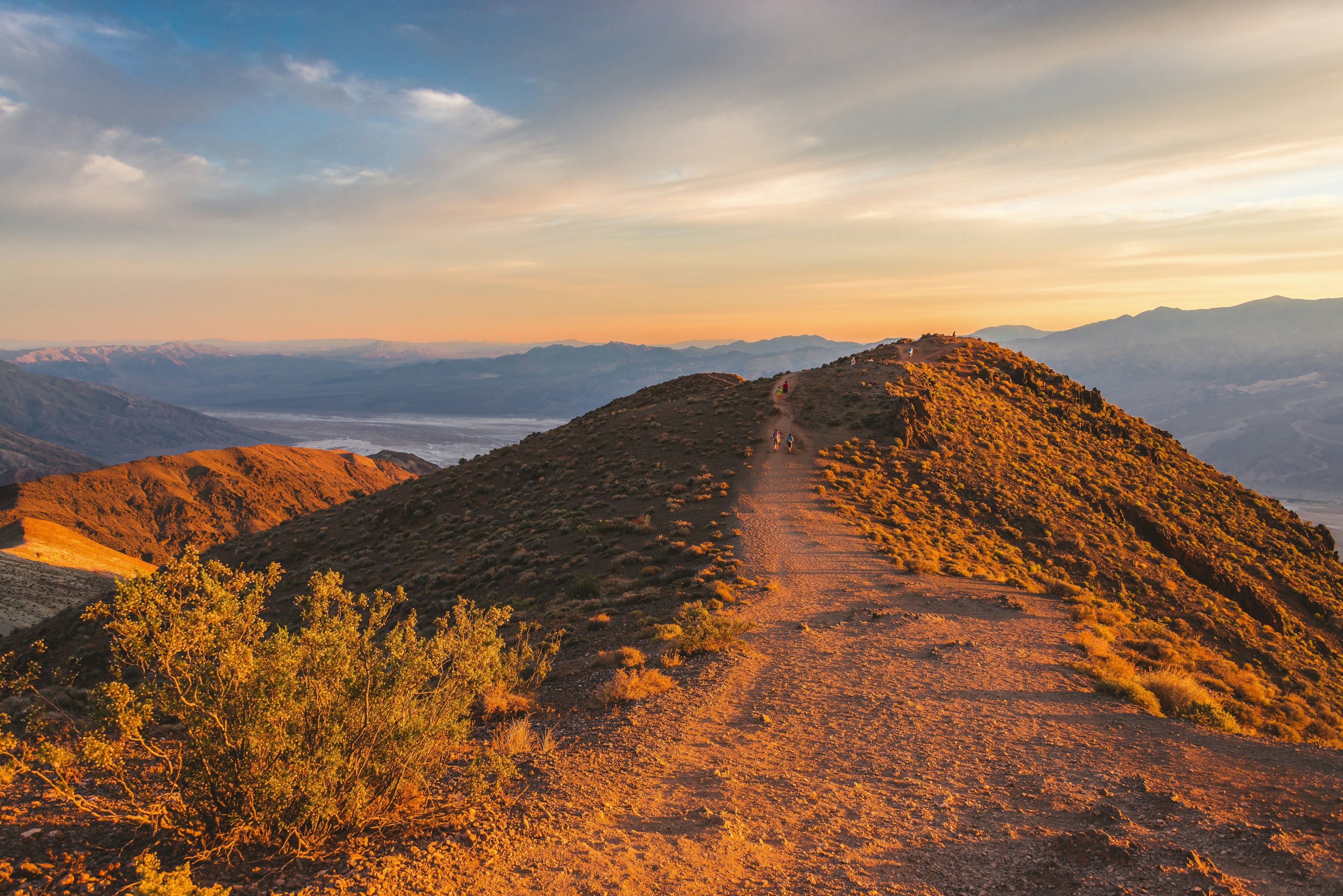 Hikers follow a dusty path up to a viewpoint from a peak