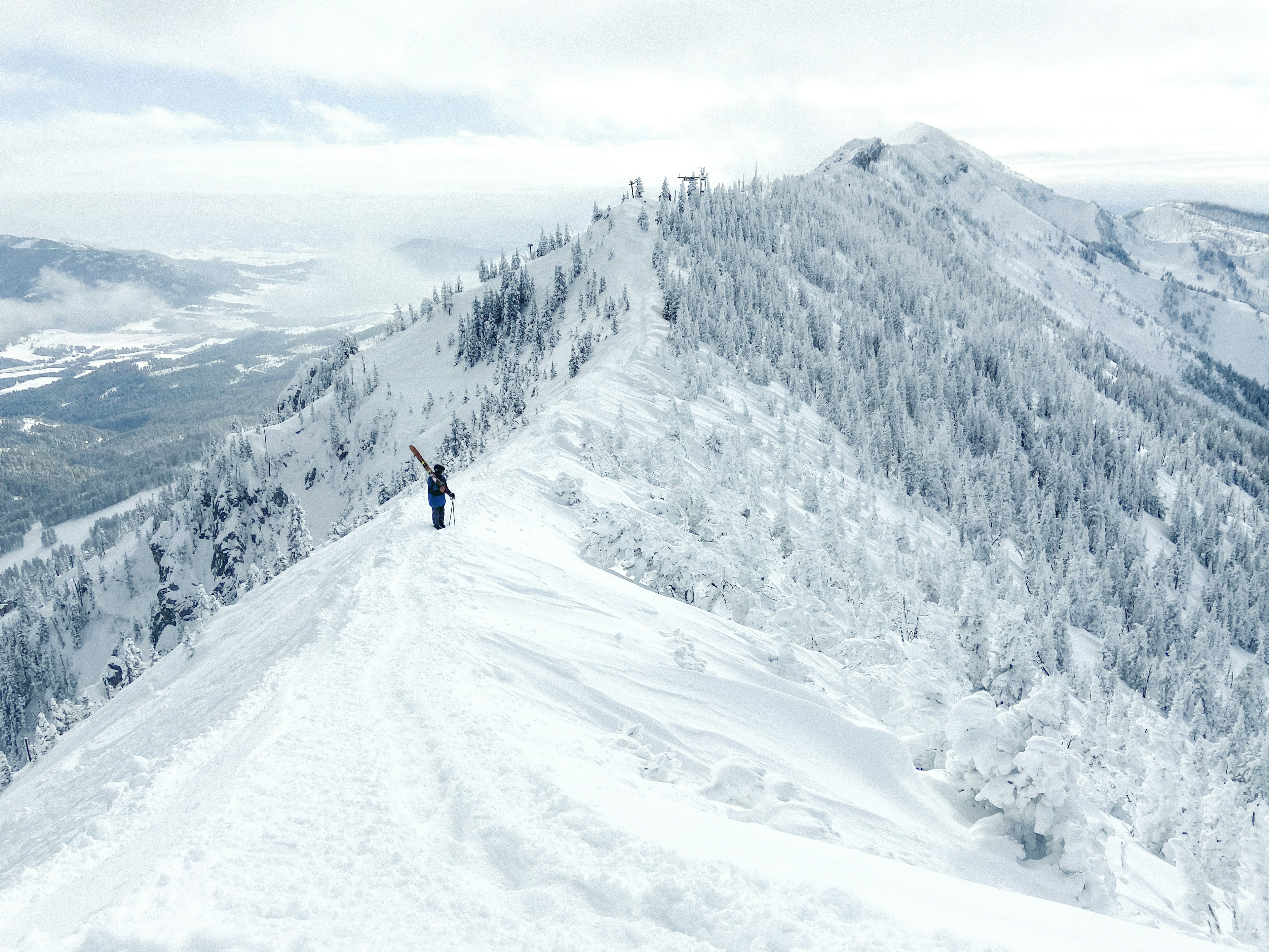 A skiier stands at the top of a snowy ridge looking out over the snow-covered slopes