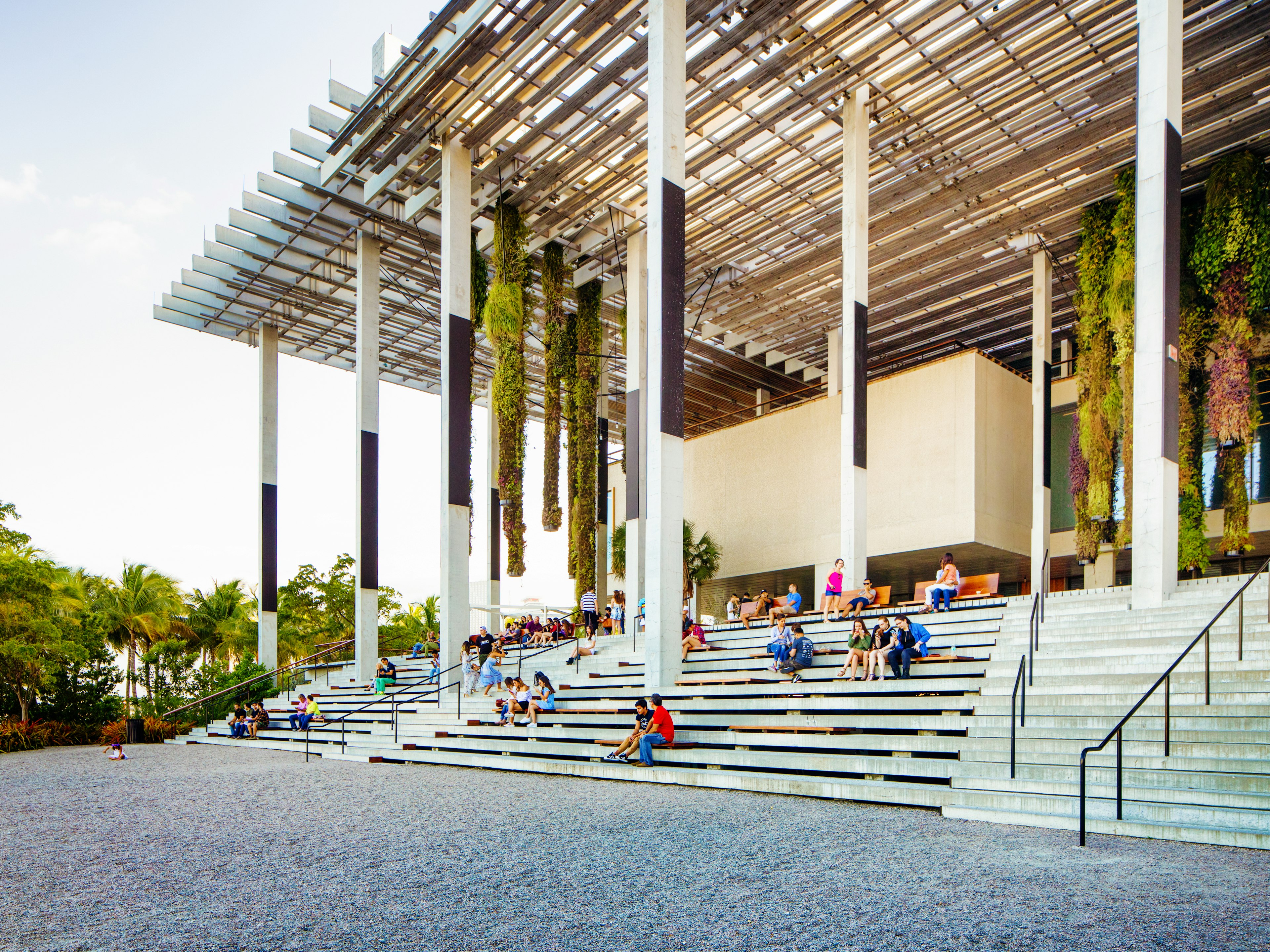 People sitting and climbing on stairs up to a museum with hanging plants coming down from its slatted awning