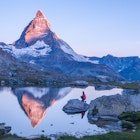 Early morning dawn scene of sunrise on the Matterhorn Mountain reflecting pink in the lake with male man on rock with red down jacket with clear blue sky Gornergrat Zermatt Matterhorn Europe