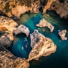 An aerial view over people stand-up paddle boarding amid golden sandstone cliffs and sea stacks, lapped by turquoise water.