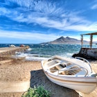 A white boat moored on a beach, with a jetty to the left and a stone structure to the right; out to sea is a dramatic peaked island.