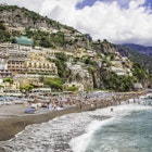 A view of the beach in Positano, on the Amalfi Coast. The beach is crowded with people, and colourful buildings climb the rocky hillside behind it.