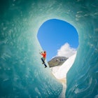 A solo male ice climber working his way up on an ice wall in a glacier. Lifestyle image created from the inside of an ice cave.