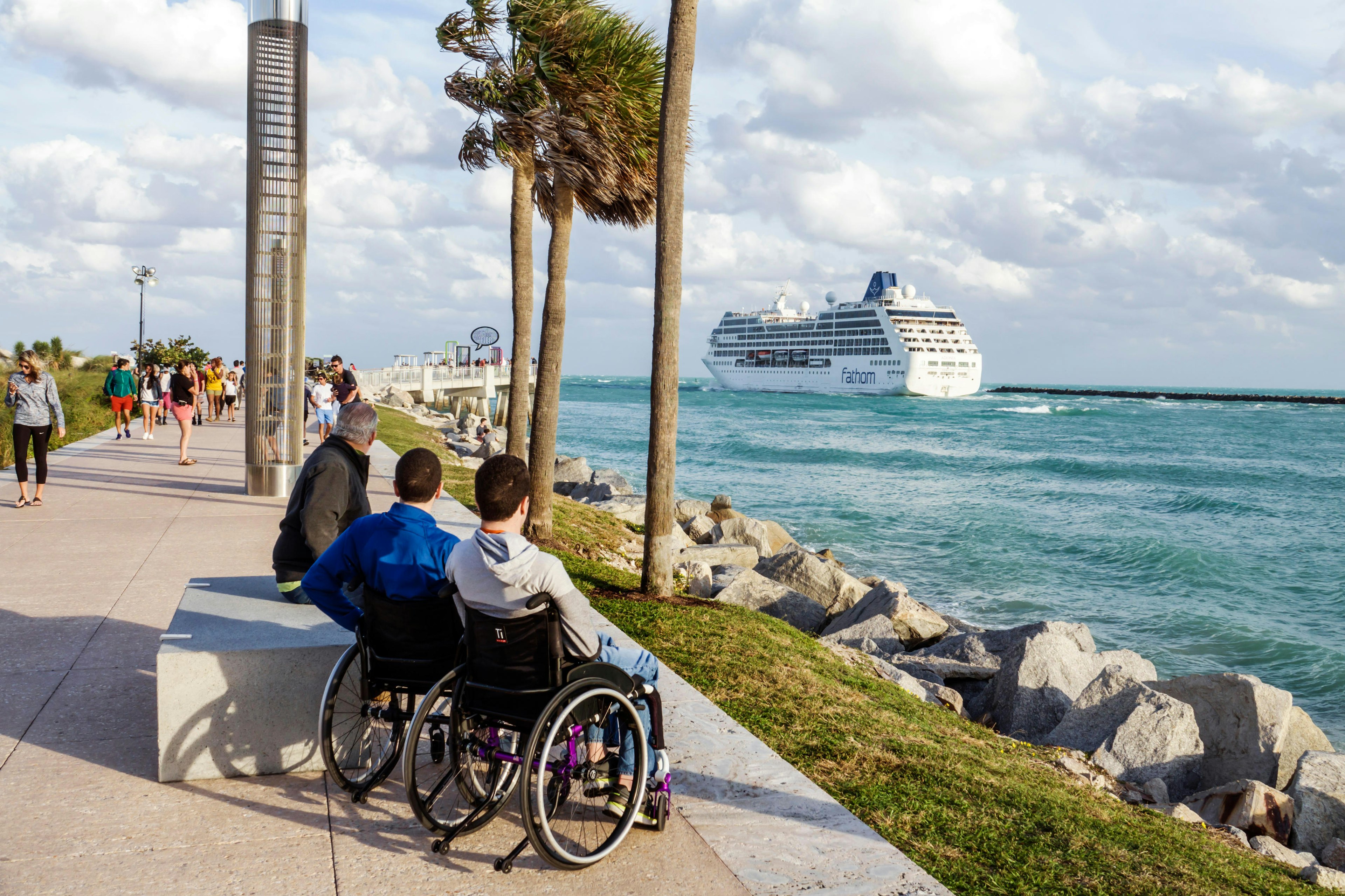 Three people, two in wheelchairs, are on the boardwalk in Miami and looking out to sea, where there's a huge cruise ship