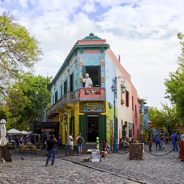 Buenos Aires, Argentina - April 15, 2015: The main square on of the Camanito in the La Boca neighborhood of Buenos Aires features brightly colored buildings and cobblestone streets that are a popular tourist destination. Tourists can be seen surrounding the most recognizable building the the neighborhood at the center of the square. The area is a popular destination for watching tango dancers in the street, shopping for souvenirs handicrafts made by local artisans and restaurants. It is the oldest neighborhood in Buenos Aires and is located at the mouth of the port, which gives it its namesake.