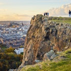 Group of people walking along cliff edge looking at city views. Edinburgh Castle in the distance