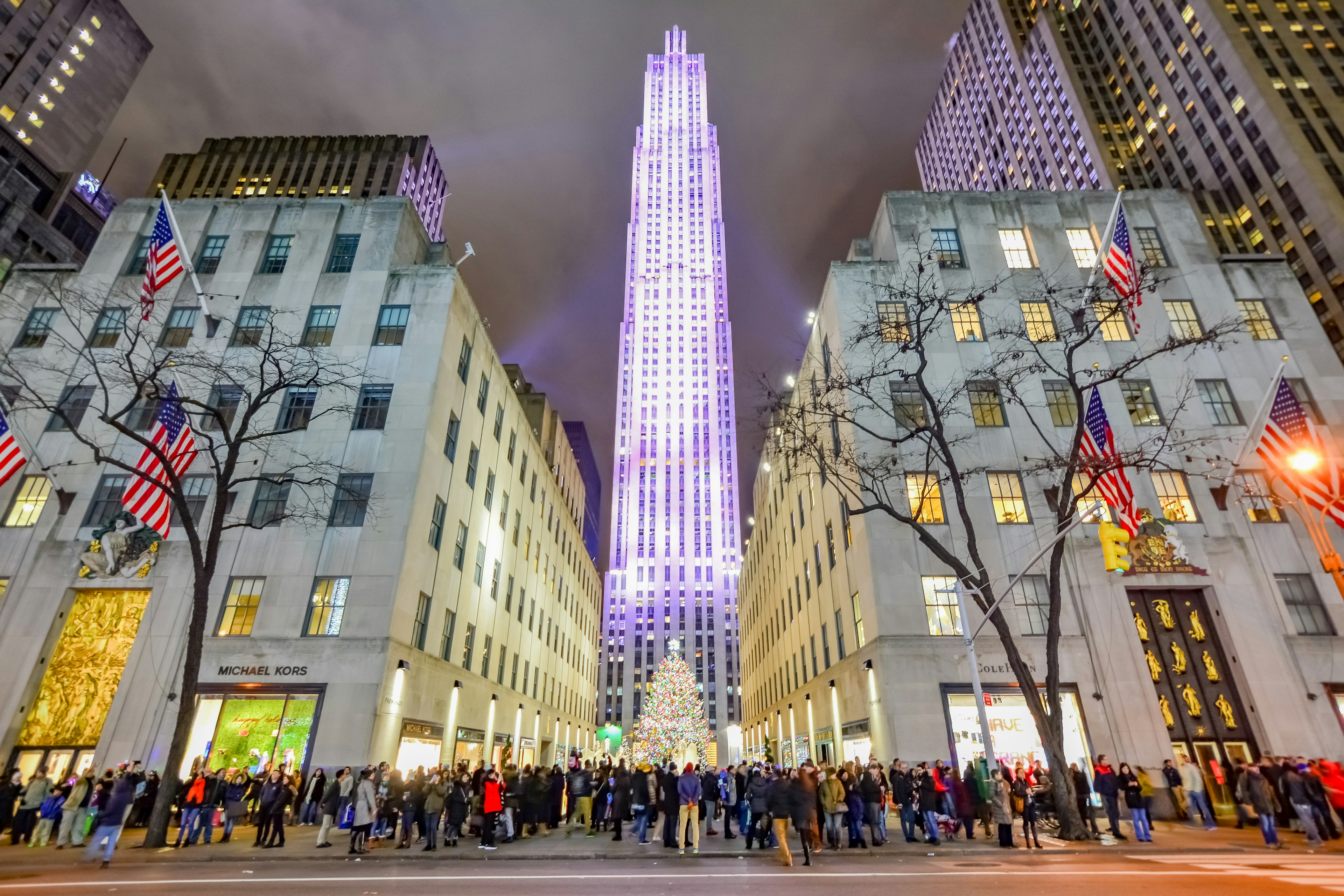 Crowds of people at the foot of a tall tower lit at Christmas time. A tall colorful tree stands in front of it